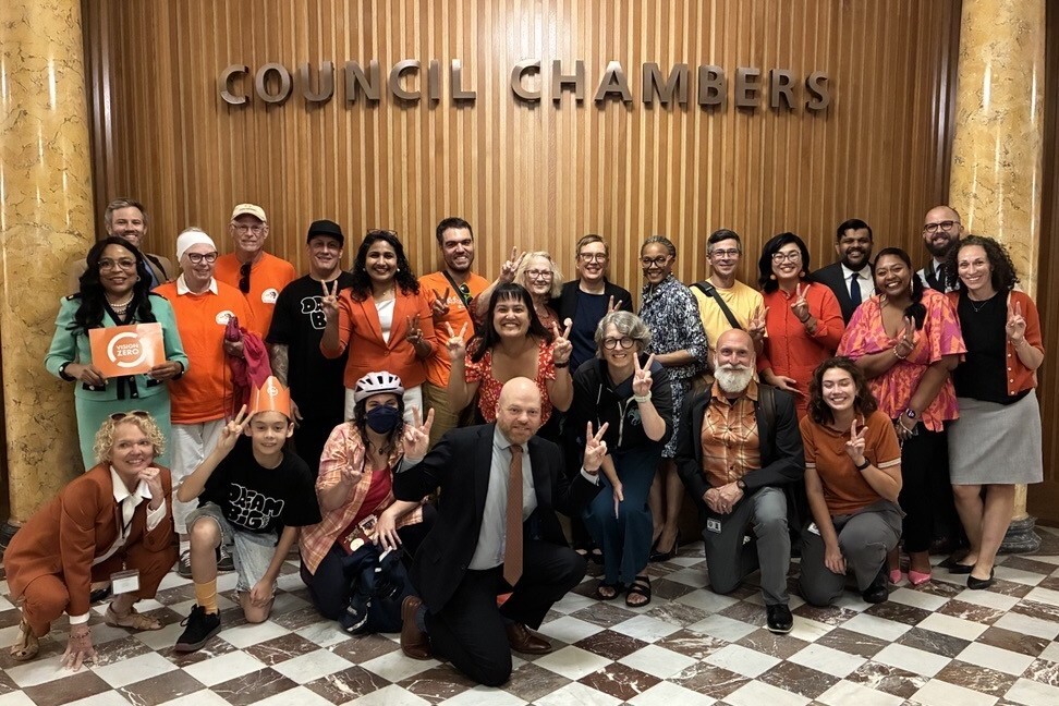 Group of smiling people, many wearing bright orange, standing in front of the Council Chambers sign at Portland City Hall