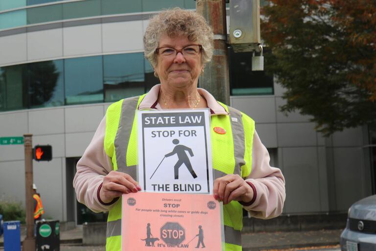 An older individual wears a reflective safety vest and holds a black and white 8.5x11 sign that reads "State law stop for the blind."