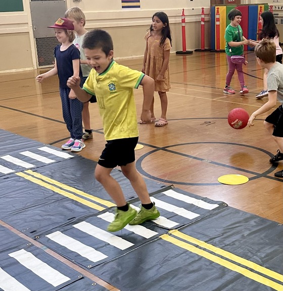 Seven elementary school-aged kids practice safely walking across a street in their gym with mock a street on the ground.