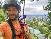 Wes Roberts wears a helmet while taking a selfie while roped and harnessed while hanging from a tall tree