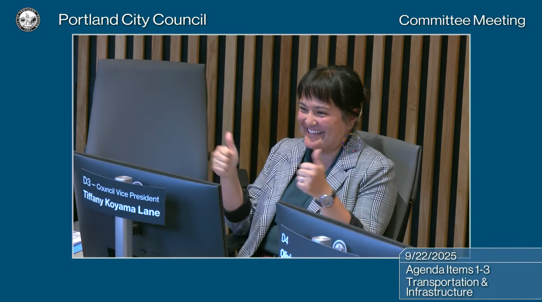 Councilor Koyama Lane, smiling and giving a double thumbs-up gesture from the City Council dais