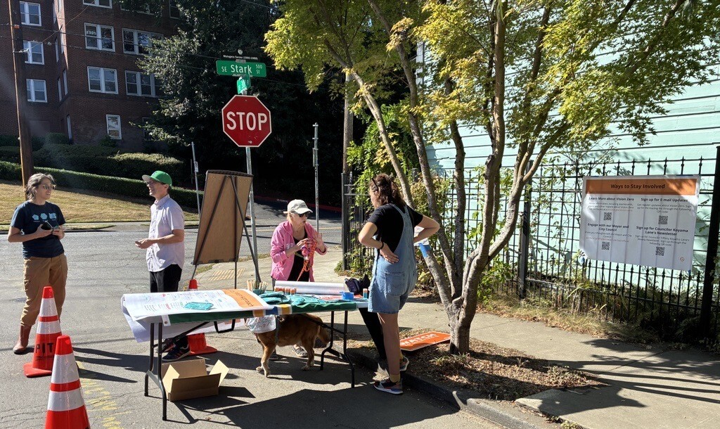 Four people outside talking in pairs outside on a sunny day, in an area in the street marked by orange traffic cones