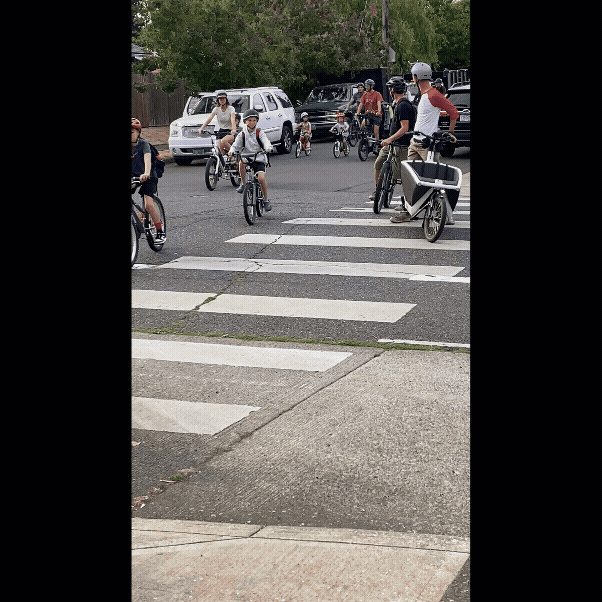 More than a dozen kids and adults on bikes approach a marked crosswalk on their way to school.