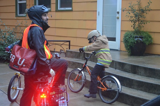 An adult and two kids wait next to a porch in rain gear with reflective elements, including bike lights.