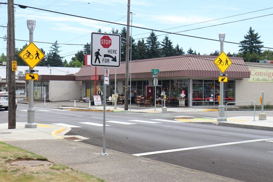 New crosswalk improvements at an intersection next to a consignment store includes lighting, flashing beacons, and signage.