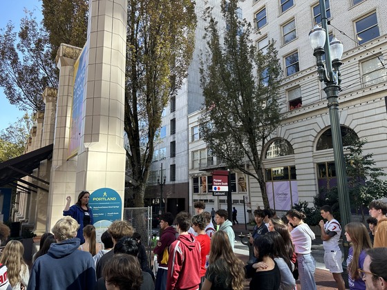 More than 30 high school students gather at downtown plaza on a sunny day to listen to an adult speaker.