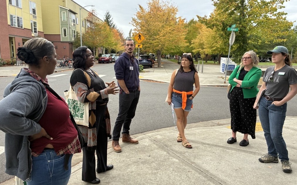 Group of people standing in a circle on a sidewalk while listening to a representative of Home Forward speaking, with trees in the background