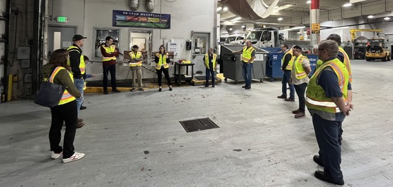 Group of people in yellow-green safety vests, standing in a circle listening to a person speak,inside a garage with large vehicles in the background.