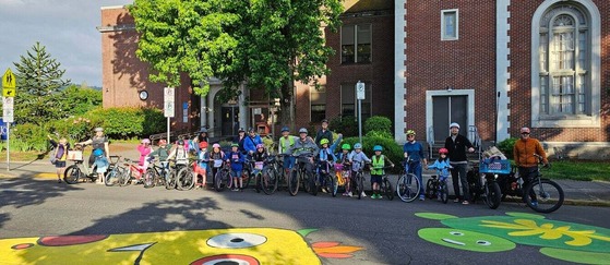 About 24 kids and adults stand in a line parallel to a neighborhood street mural on their bikes in front of a brick building.