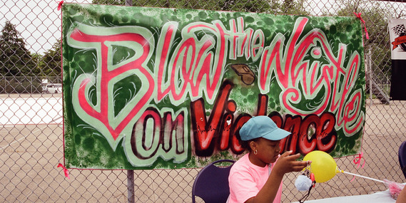 A girl sits in front of a hand-painted sign that reads "Blow the whistle on violence"