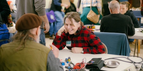 Repair Cafe shows a woman smiling at a table while talking with someone fixing her appliance