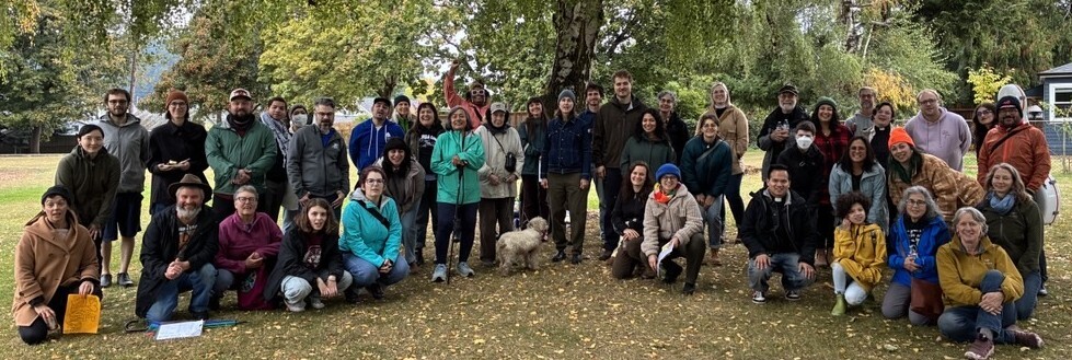 Large group of people facing camera and smiling. Picture is in a park with trees, with some yellow fallen leaves on the grass.