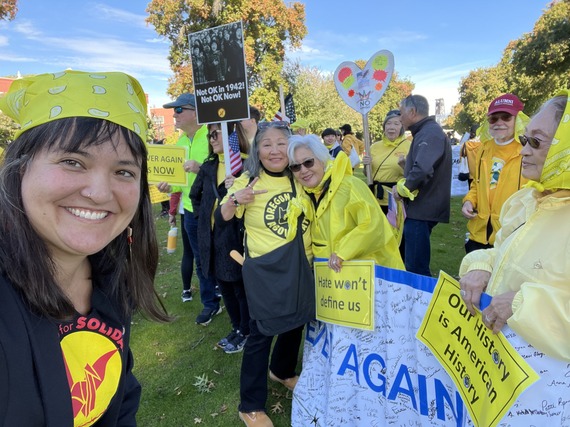 Group of people, wearing yellow, holding a banner that says "Never Again" and various signs including one that says "Our History is American History"