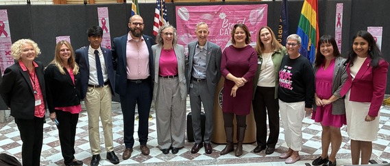 Line of people, most wearing pink, facing the camera. Behind them are Portland, US and Pride flags, as well as a lectern.