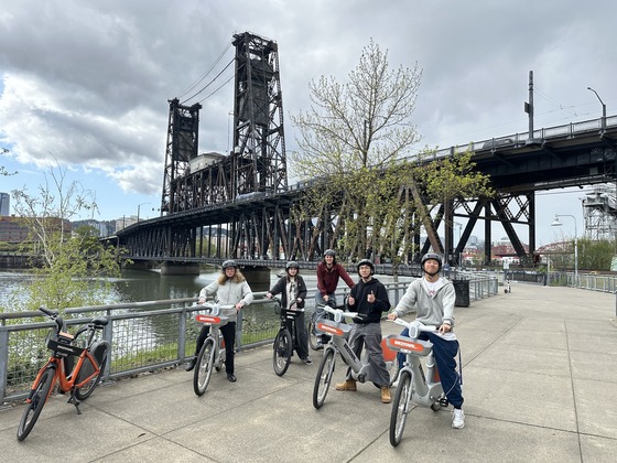 Five teenagers ride BIKETOWN bike-share e-bikes on Portland's waterfront near the Steel Bridge. 