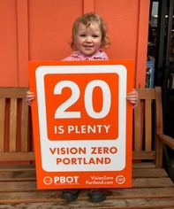A young child wearing a pink rain jacket stands on a wood bench holding an orange "20 is plenty" vertical yard sign.