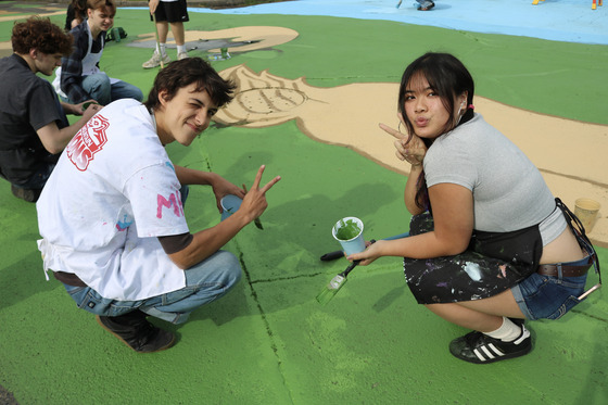 Two high school students squatting while painting a colorful street mural in a neighborhood intersection.