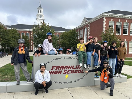 One adult teacher and 12 high school students with bike helmets on stand around Franklin High School's permanent sign.