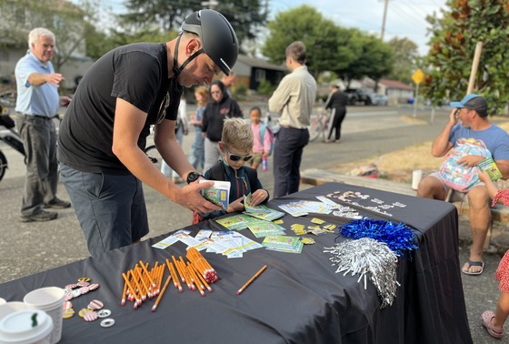 An adult helps a young child take a small comic book from a table outside a school on a morning. 