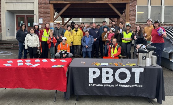 More than 20 adults and kid stand under a small bike shelter outside a school, behind a table with a PBOT logo. 