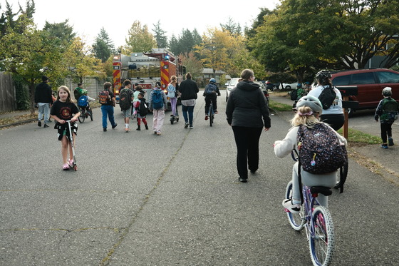 About 15 adults and kids walk to school through a neighborhood behind a red fire engine. 