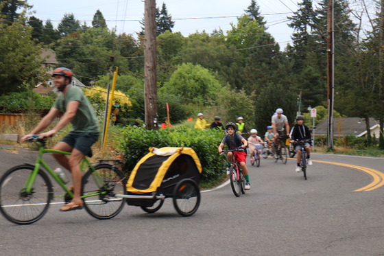 About eight adults and kids turn the corner on a neighborhood road on a bike ride to school in the morning. 