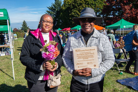 Two smiling people stand outside, one holding fresh flowers and the other holding a plaque award.