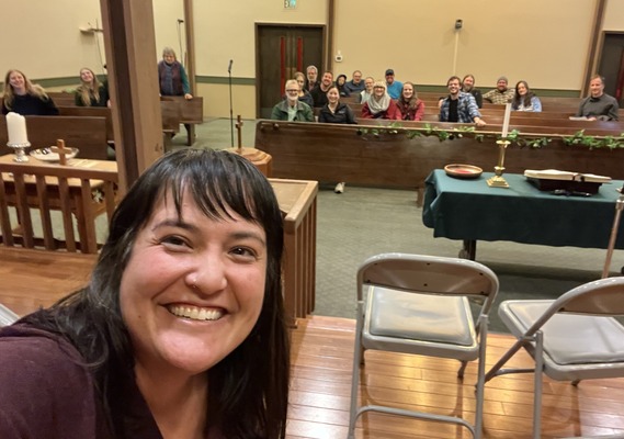 Councilor Koyama Lane on left, smiling at camera, in front of several rows of smiling people sitting on wooden pews and looking at the camera