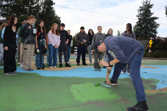 About 12 teenagers standing on a street mural huddle around an adult instructor demonstrates painting technoque.