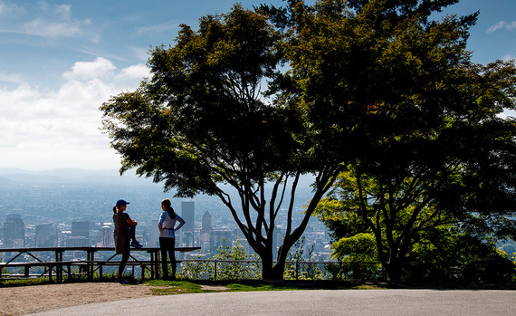 Two people are silhouetted with a view of downtown Portland in the background and trees next to them