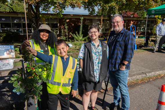 Photo shows four people of diverse gender, age and ethnicity smiling at camera while displaying a young tree ready to be planted