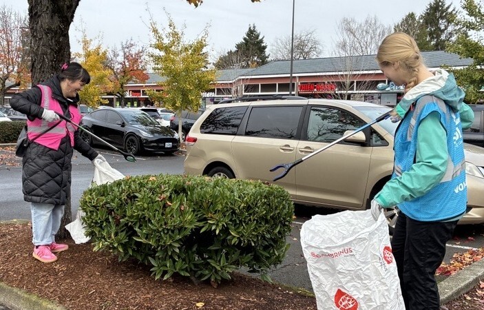 Two people using graspers to collect trash into plastic bags in a parking lot