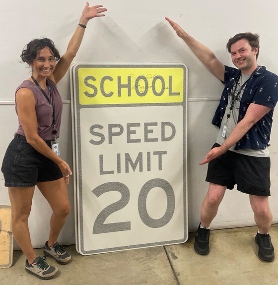 Two adults extend their arms to frame a large school 20 mph speed limit sign. 