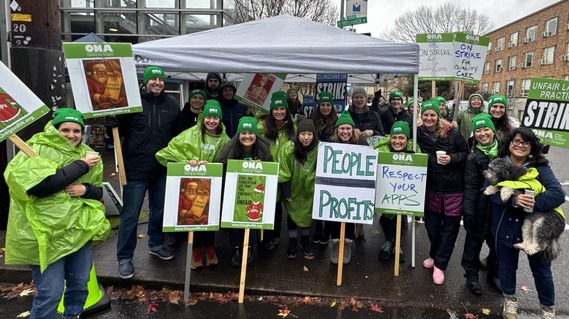 group of people, many wearing green rain ponchos, holding signs that include "on strike for quality care, "people over profits" and "ONA"