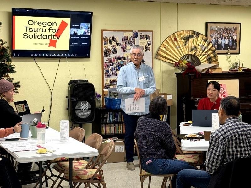 Group of people in room, some sitting at tables and one person standing and presenting next to a monitor that reads Oregon Tsuru for Solidarity