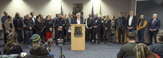 Governor Tina Kotek stands at a podium during a press conference while Mayor Keith Wilson and many other community leaders stand next to her