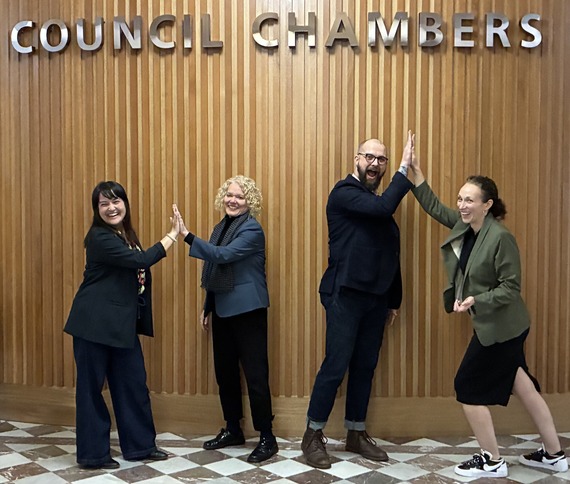 Councilor Koyama Lane and Clark high-fiving and Councilor Dunphy and Pirtle-Guiney high-fiving in front of the wood Council Chambers backdrop