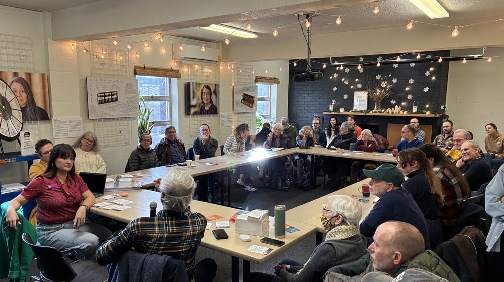 large group of people sitting around a ring of tables, listening to a speaker