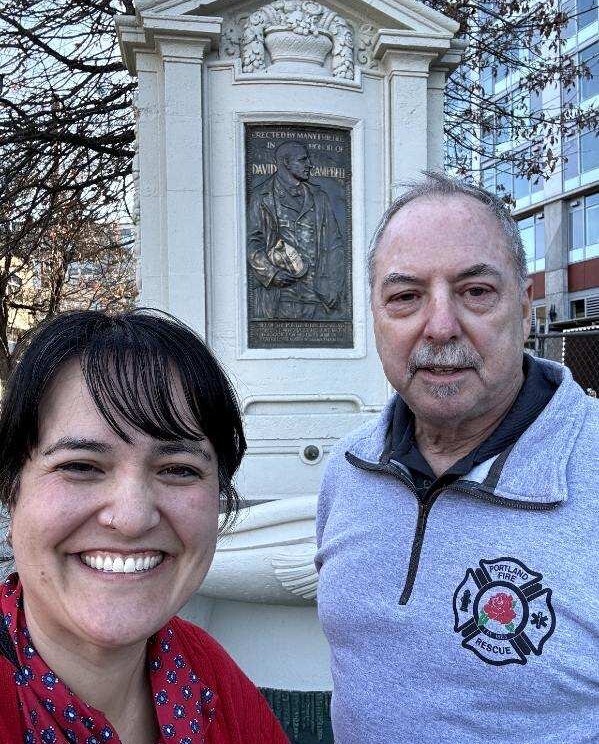 Picture of Councilor Tiffany Koyama Lane and Don Porth, smiling in front of stone and brass memorial to fallen firefighter David Campbell