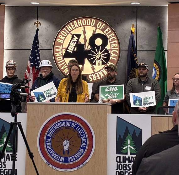 Six people, some in hard hats and other work uniforms, holding signs about Climate Jobs Oregon and Clean Energy, behind a person at lectern