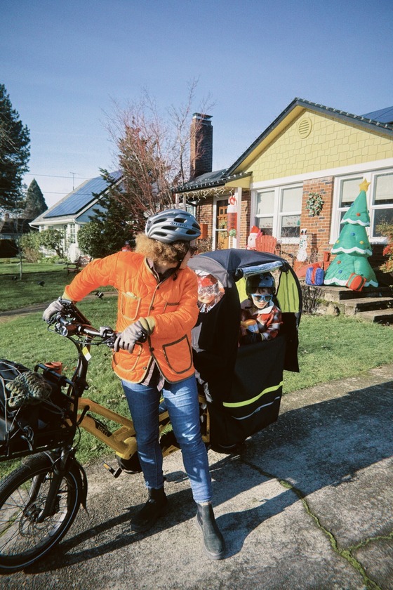 An adult stands with an e-bike carrying a young child in the driveway of a home decorated for Christmas.