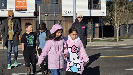 Four elementary school students and two adults walk across a marked crosswalk together on a cold day.