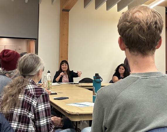 Councilor Koyama Lane holding microphone, sitting with group of people around a rectangle of tables
