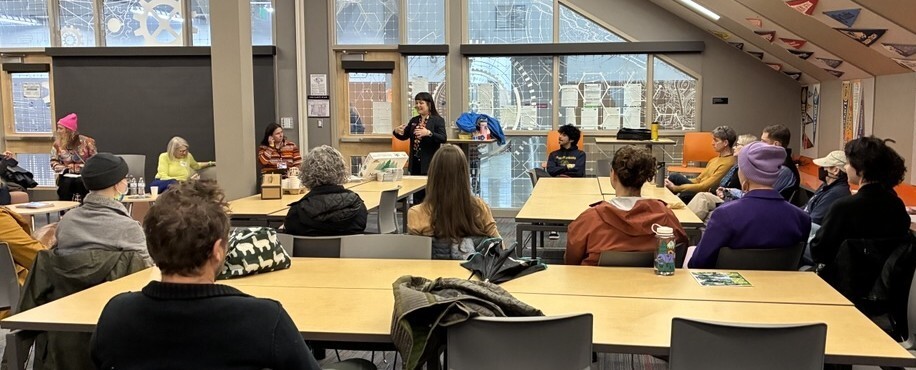 Room full of people sitting at tables, watching Councilor Koyama Lane, standing and holding a microphone, speak