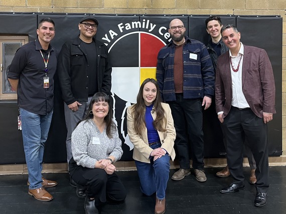 Group of people, some standing and some kneeling, in front of hanging gymnasium mat printed with NAYA Family Center logo