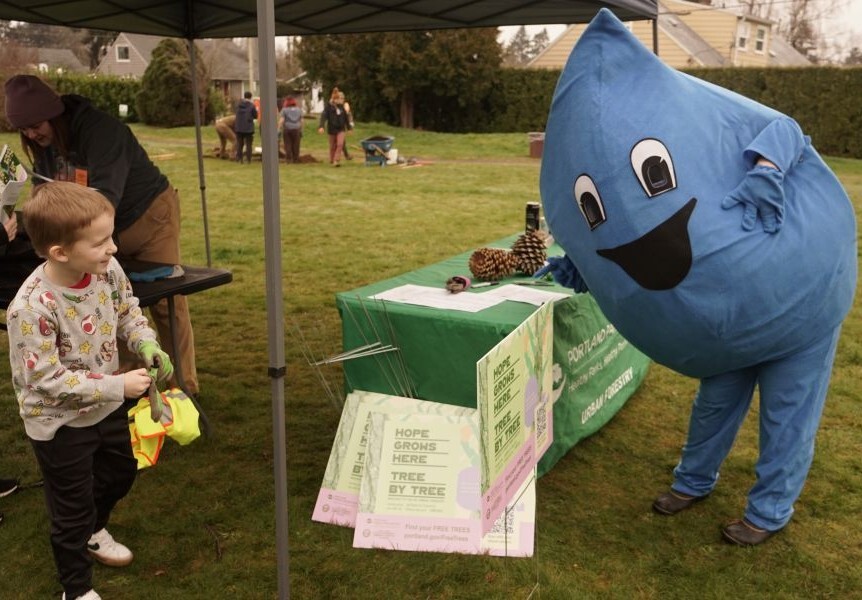 PPR - Water drop mascot talks to young child in front of a green table and yard signs