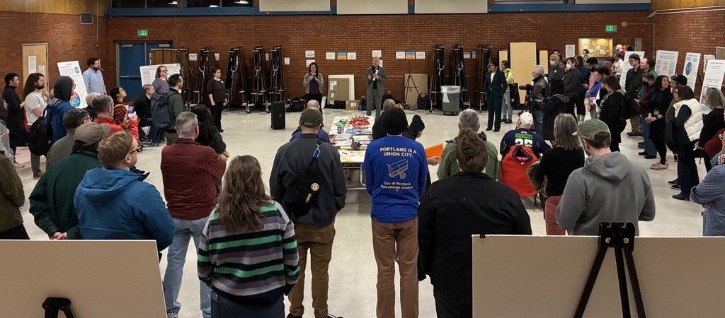 group of people standing in a circle in a large room, watching a person speak at a microphone. Large posters on easels around the room