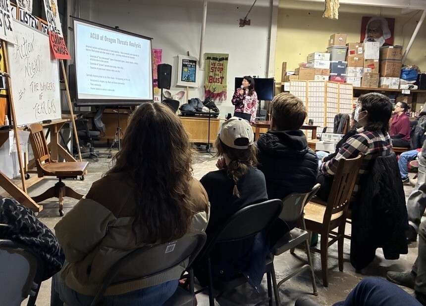 Speaker holding microphone and looking at projector screen that reads "ACLU of Oregon Threats Analysis," rows of people in seats watching
