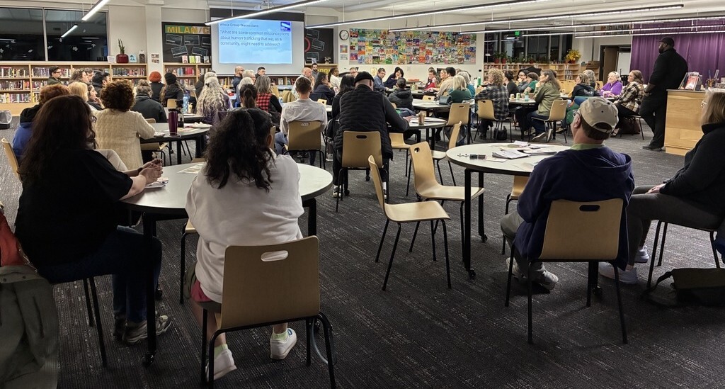 Group of people sitting at tables and chairs in McDaniel High School's library, with projector screen in background
