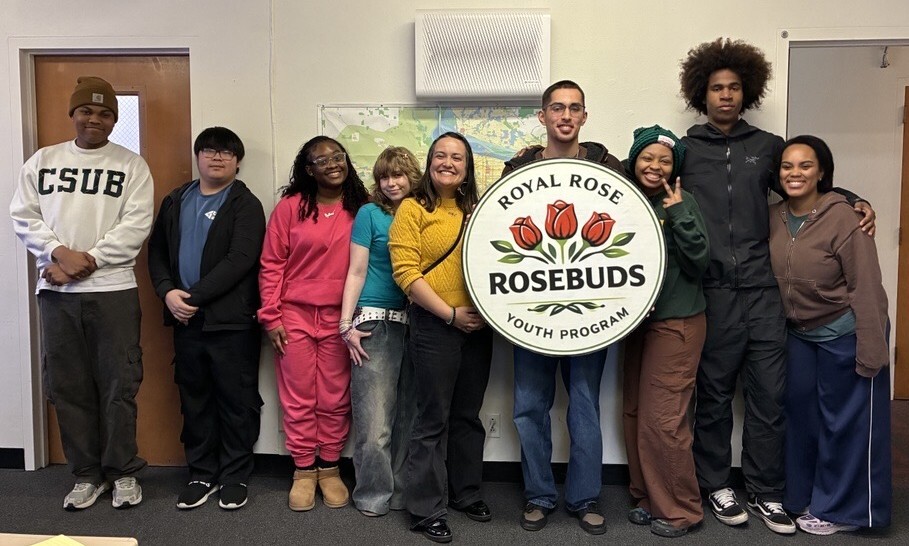 Group of nine people standing and smiling, holding a round sign that reads "Royal Rose Rosebuds Youth Program" with three roses in the center